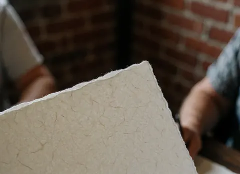 Feuille de papier texturé aux bords déchirés tenue par des mains devant un mur de briques rouges, évoquant l'artisanat et la qualité.