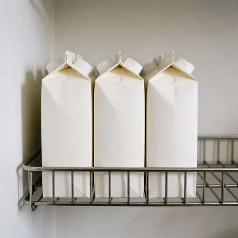 Trois cartons de lait blanc sans marque posés sur une étagère métallique devant un mur blanc avec un éclairage doux, idéal pour un minimarket ou une épicerie.