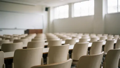 Vue d'une salle de cours déserte comportant des chaises beige alignées, un tableau noir et un pupitre, éclairée par la lumière naturelle entrant par les fenêtres.