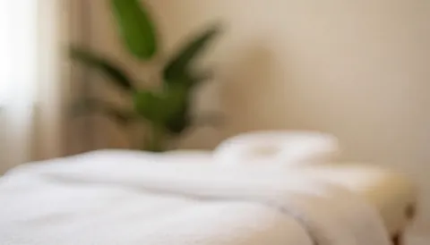 Vue d'une salle de massage calme avec une table de massage, des serviettes blanches et une plante verte en arrière-plan flou.