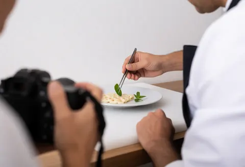 Un styliste culinaire place délicatement une feuille d'herbe sur un plat à l'aide de pinces, dans une cuisine lumineuse aux murs blancs.
