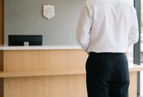Vue arrière d'une personne debout dans une réception moderne avec bureau en bois clair, écran noir et mur gris.