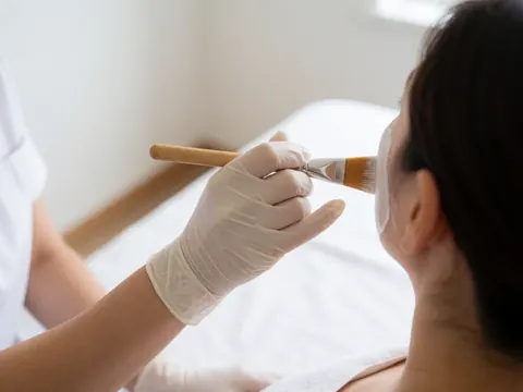 Professionnel portant un gant blanc applique un masque facial avec une brosse en bois sur un client, fond blanc, lumière naturelle, soin esthétique.