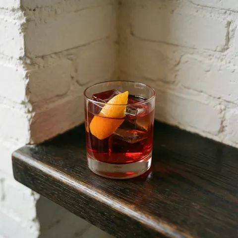 Un cocktail negroni classique servi dans un petit verre avec des glaçons et une tranche d'orange sur un fond sombre.
