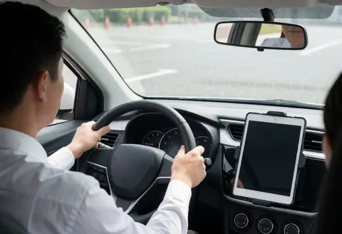 Un instructeur d'auto-école tient le volant d'une voiture blanche dans un terrain d'entraînement, avec une tablette posée sur le tableau de bord et des cônes en arrière-plan sous la lumière du soleil.