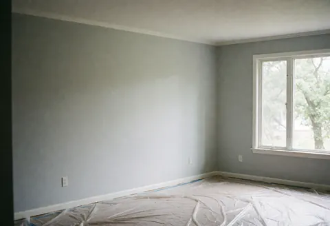Intérieur d'une pièce rénovée avec murs gris, sol protégé par du plastique et lumière naturelle entrant par une fenêtre.