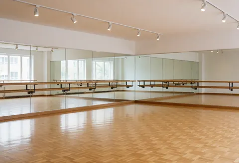 Studio de danse déserté présentant un sol en parquet poli, des barres de ballet et des miroirs éclairés par la lumière du plafond.