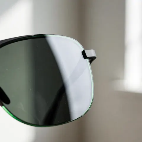 Détail rapproché d'une lentille de lunettes de soleil présentant un fini vert et des gouttelettes d'eau, illustrant la technologie hydrophobe sur fond flou aux tons neutres.