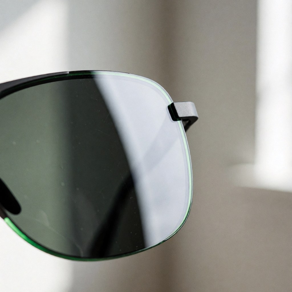 Détail rapproché d'une lentille de lunettes de soleil présentant un fini vert et des gouttelettes d'eau, illustrant la technologie hydrophobe sur fond flou aux tons neutres.