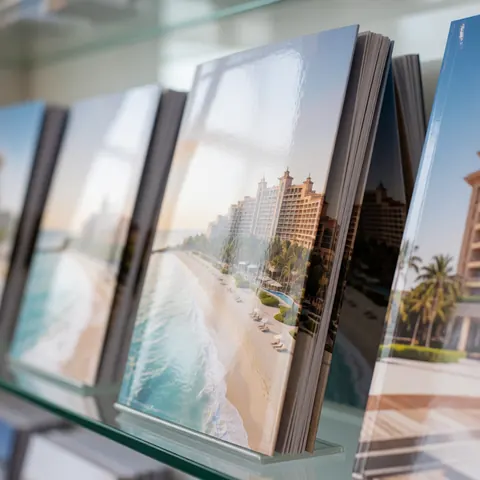 Une brochure publicitaire pour un hôtel de luxe posée sur une étagère en verre près d'une fenêtre avec vue sur la plage et les palmiers.