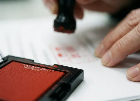 Macro d'une main appliquant un tampon encreur rouge sur un papier blanc, avec un arrière-plan flou, évoquant la comptabilité et l'administration.