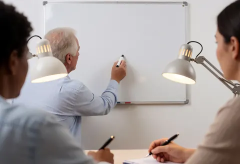 Un tuteur écrit sur un tableau blanc pendant que deux étudiants prennent des notes avec des stylos sous l'éclairage de lampes de bureau dans une classe réaliste.