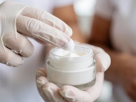 Des mains portant des gants blancs saisissent une portion de crème hydratante dans un bocal en verre sur un fond flou et professionnel.
