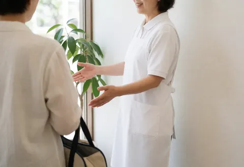 Un thérapeute en blanc accueille un client portant un sac dans l'entrée lumineuse d'un centre de santé, entouré de plantes.