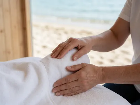 Gros plan des mains d'un thérapeute pratiquant un massage sur un pied posé sur une serviette blanche, avec une vue sur la mer et le bois en arrière-plan.