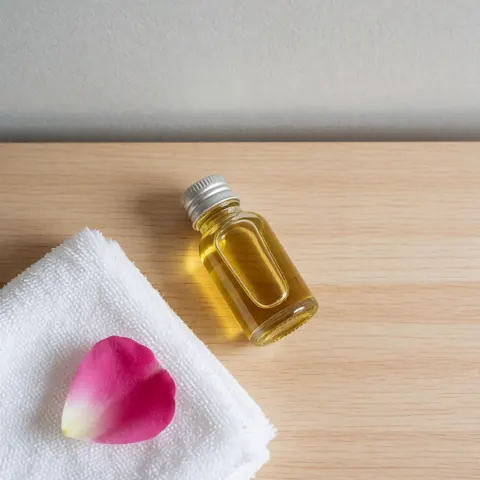 Bouteille d'huile faciale à la rose posée sur une table en bois clair, accompagnée d'une serviette blanche et d'un pétale de rose, éclairée par la lumière naturelle.