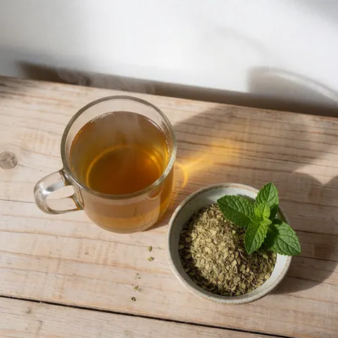 Une tasse de yerba mate fumante posée sur une table en bois clair, accompagnée d'une petite tasse contenant des feuilles sèches et de la menta fraîche.