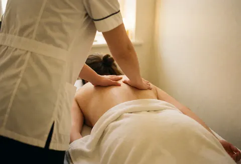 Un thérapeute en uniforme blanc effectue un massage dorsal sur un client recouvert d'un tissu blanc, dans une pièce aux murs chauds et à la lumière douce.