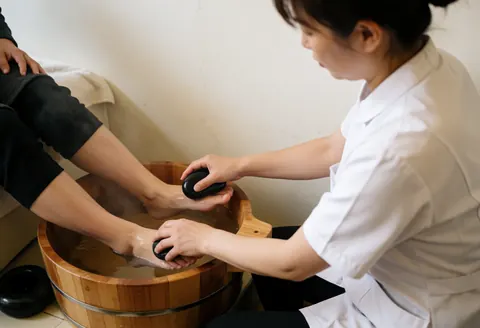 Un thérapeute en train de masser les pieds d'un client avec des pierres de basalte chaudes dans un cadre relaxant et professionnel.
