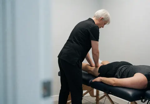 Un thérapeute professionnel réalise un massage sur la jambe d'un client allongé sur une table de massage dans un espace lumineux et neutre.