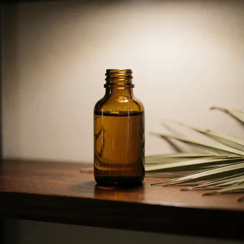 Une bouteille en verre ambrée contenant de l'huile de palme est posée sur une étagère en bois sombre, accompagnée de feuilles de palmier séchées.