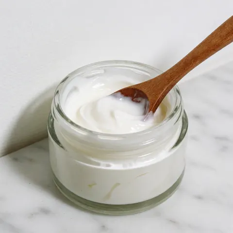 Crème blanche dans un bocal en verre avec une cuillère en bois sur une surface en marbre blanc, idéale pour les soins de la peau et le bien-être.