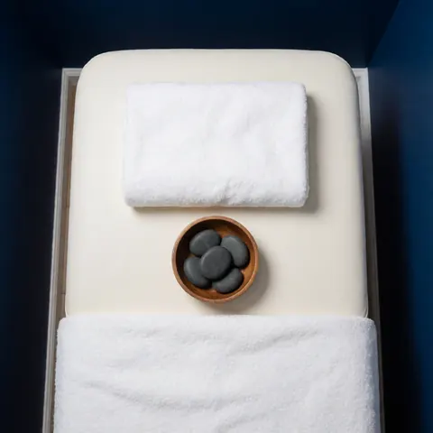 Vue de dessus d'une table de massage avec une serviette blanche, un bol en bois contenant des pierres chaudes et des outils de massage pour la thérapie.