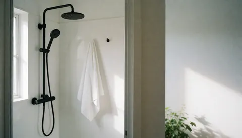 Douche au design contemporain affichant une serviette blanche suspendue, des murs blancs, une lumière naturelle entrant par une fenêtre et une plante verte.
