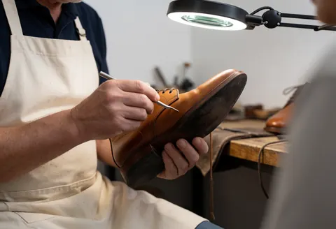 Un cordonnier portant un tablier blanc répare une chaussure en cuir marron sous un éclairage intense dans son atelier.