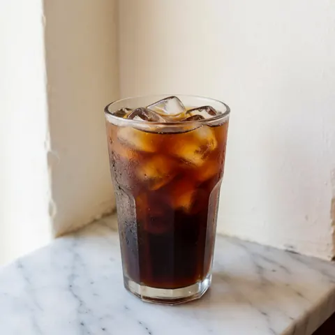 Un café glacé frais avec des glaçons est présenté dans un verre transparent posé sur une surface en marbre blanc veiné de gris.