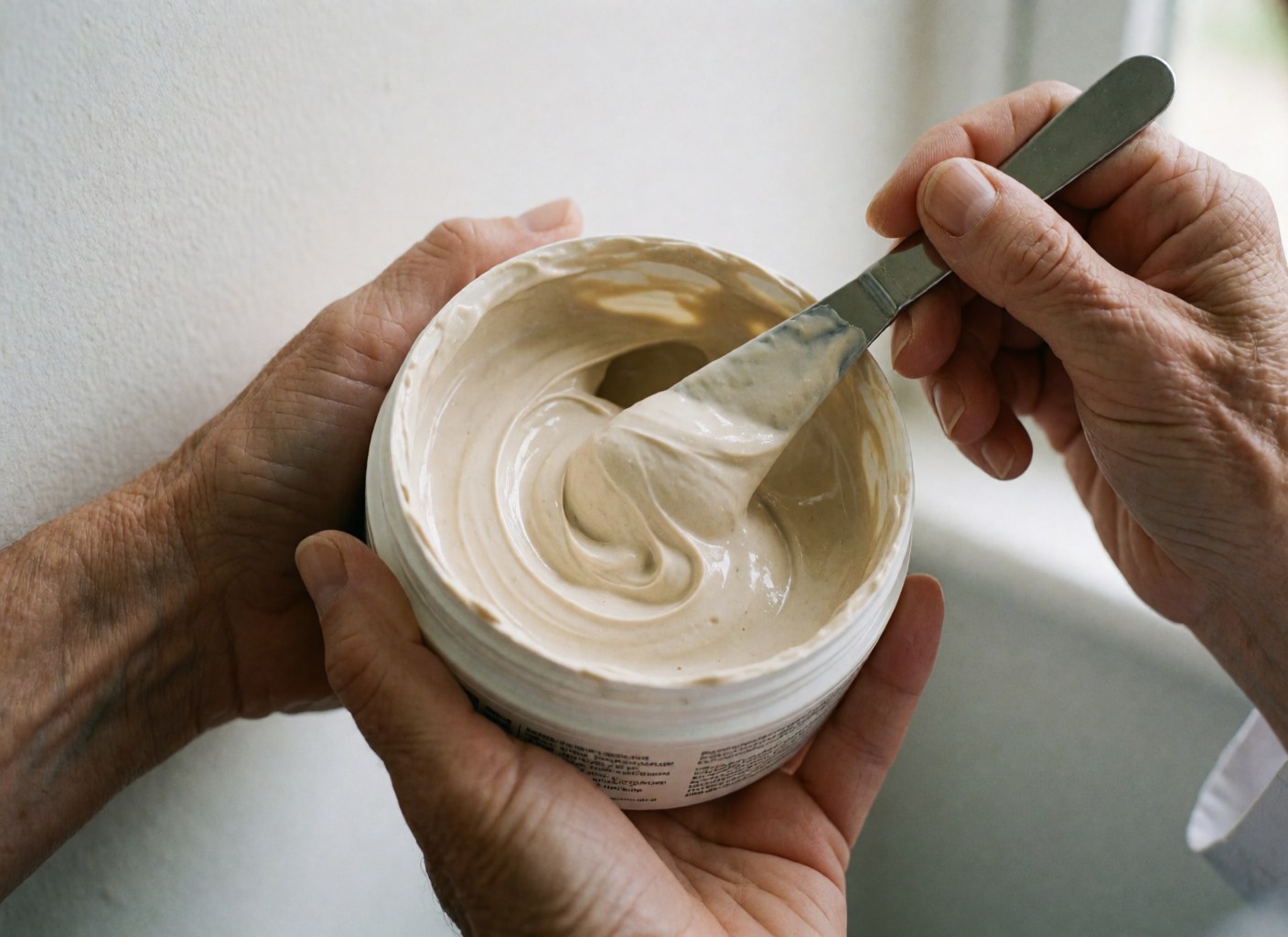 Gros plan d'un masque capillaire beige dans un pot blanc, appliqué avec une spatule métallique tenue par des mains. Lumière naturelle sur la texture crémeuse.