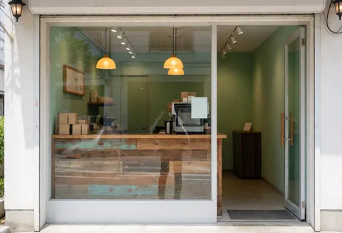 Façade d'un café avec murs verts, lumières chaudes et un comptoir en bois rustique visible par la fenêtre. L'entrée est ouverte et l'ambiance est accueillante.