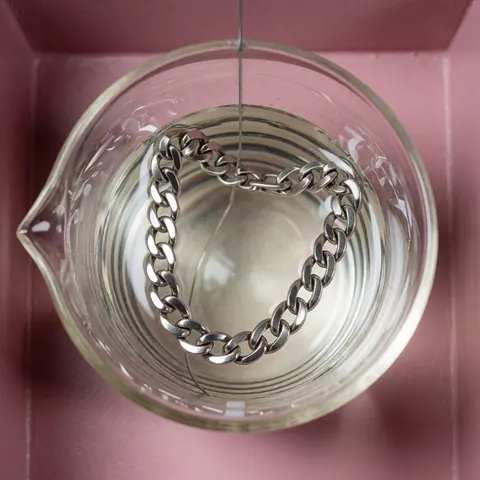 Chaîne en argent plongée dans un bécher contenant une solution électrochimique pour le processus de revêtement sur fond rose propre et technique.