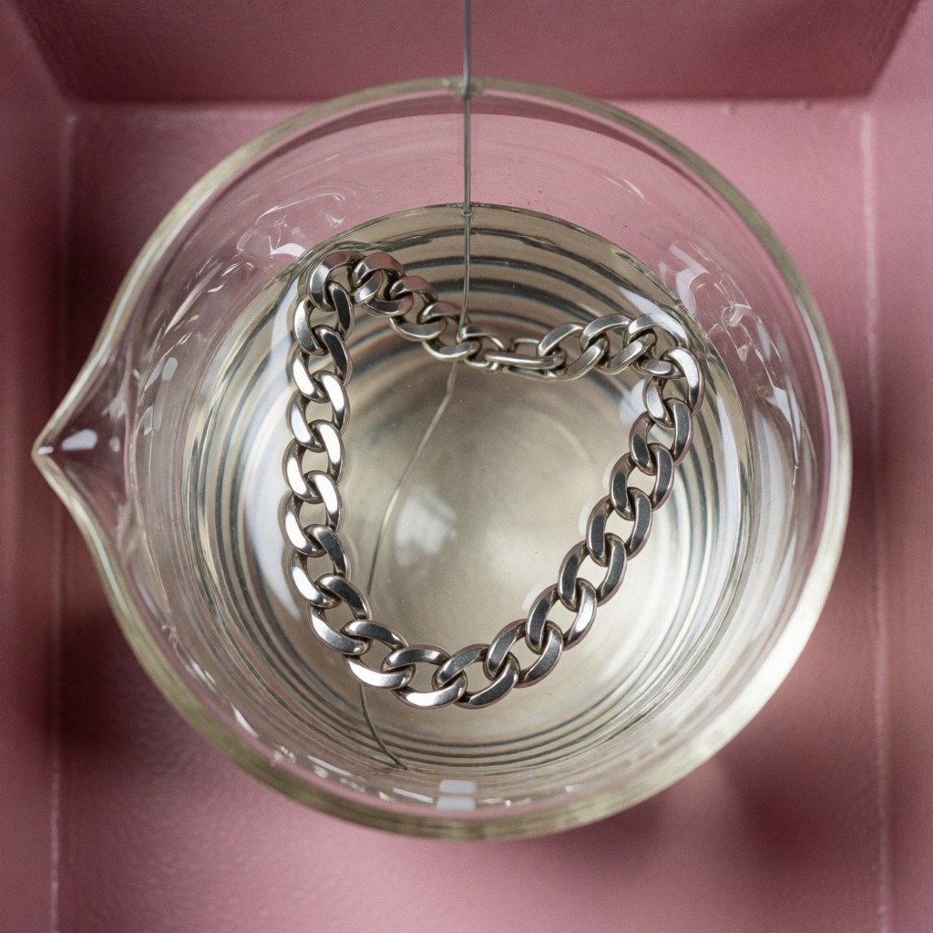 Chaîne en argent plongée dans un bécher contenant une solution électrochimique pour le processus de revêtement sur fond rose propre et technique.