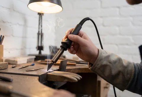 Un artisan soudeur effectue une soudure sur des joints de vitrage dans un atelier avec fumée visible.