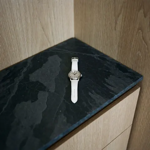 Une montre de luxe avec un cadran argenté et un bracelet blanc posée sur une surface en bois clair.