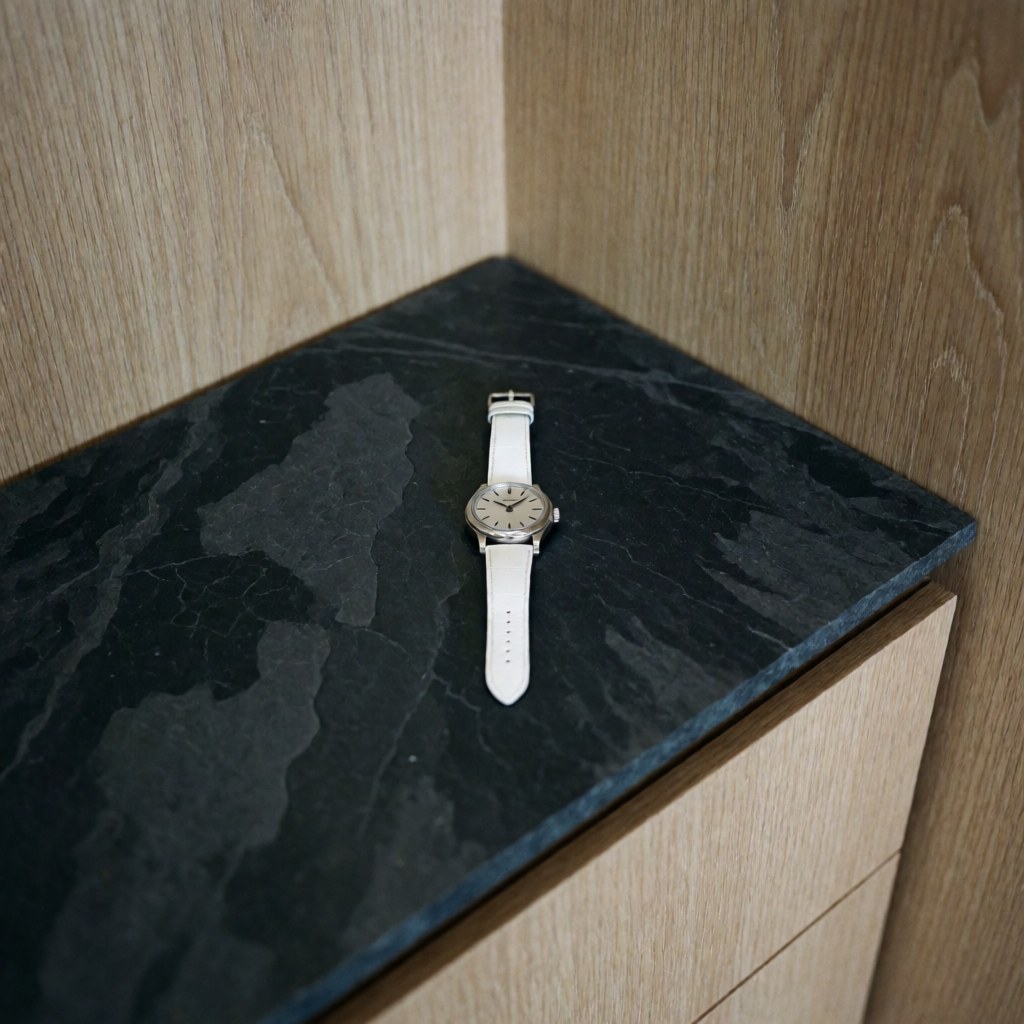 Une montre de luxe avec un cadran argenté et un bracelet blanc posée sur une surface en bois clair.