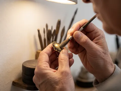Mains d'un joaillier appliquant du brillant avec un pinceau sur un broche en bronze ancien, outils de bijouterie en arrière-plan flou et lumière chaude.