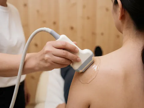Vue rapprochée d'un transducteur d'ultrasons posé sur la peau d'une épaule, avec du gel de couplage visible, dans un cadre médical professionnel.