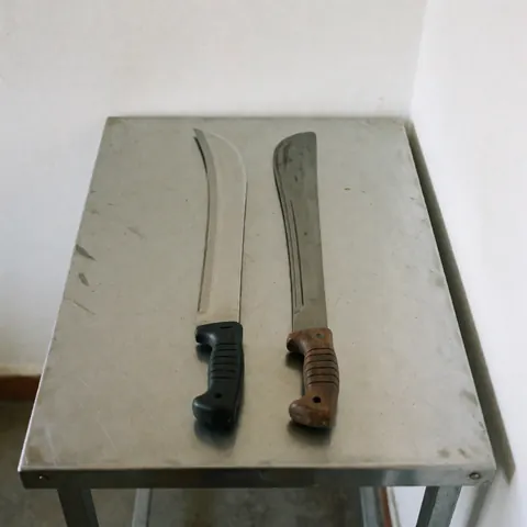 Couteau, machette et outils agricoles disposés sur une table métallique avec un fond blanc uni pour la présentation du produit.