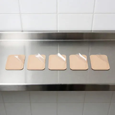 Cinq patchs transdermiques beige avec film transparent partiellement retiré, alignés sur une surface en acier inoxydable brillant, fond de carreaux blancs, scène stérile pour l'ostéopathie.