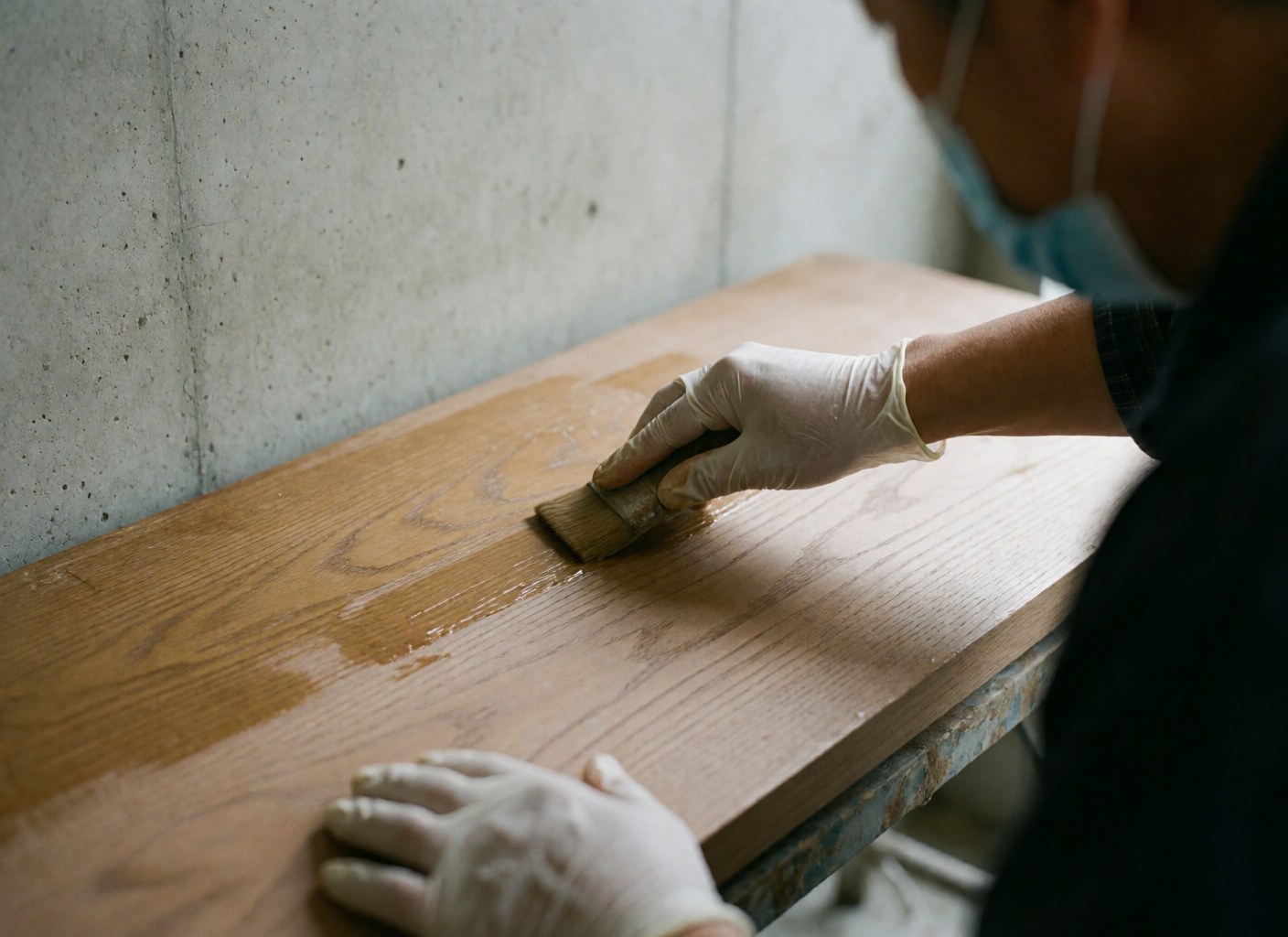Main gantée appliquant de la cire mate sur une surface en bois poli avec un pinceau, sur fond de mur en béton.