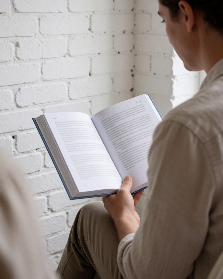 Une personne adulte lit attentivement un livre ouvert dans un intérieur calme avec un mur en briques blanches, évoquant sérénité et concentration.