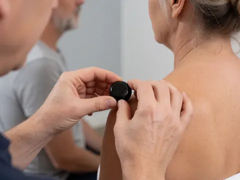 Des mains de thérapeute placent une pierre noire d'obsidienne sur l'épaule d'un client lors d'une séance de bien-être, avec un fond flou et une lumière douce.