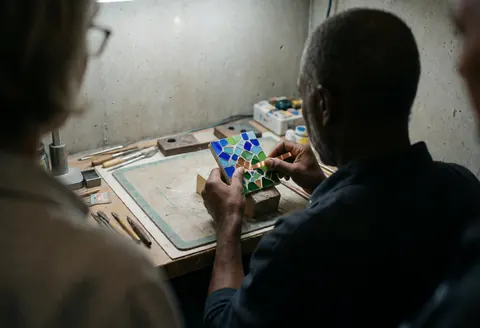 Vue arrière d'un artisan travaillant sur un panneau de marbre décoratif avec du cuivre, des outils et des matériaux dans un atelier aux murs de béton.
