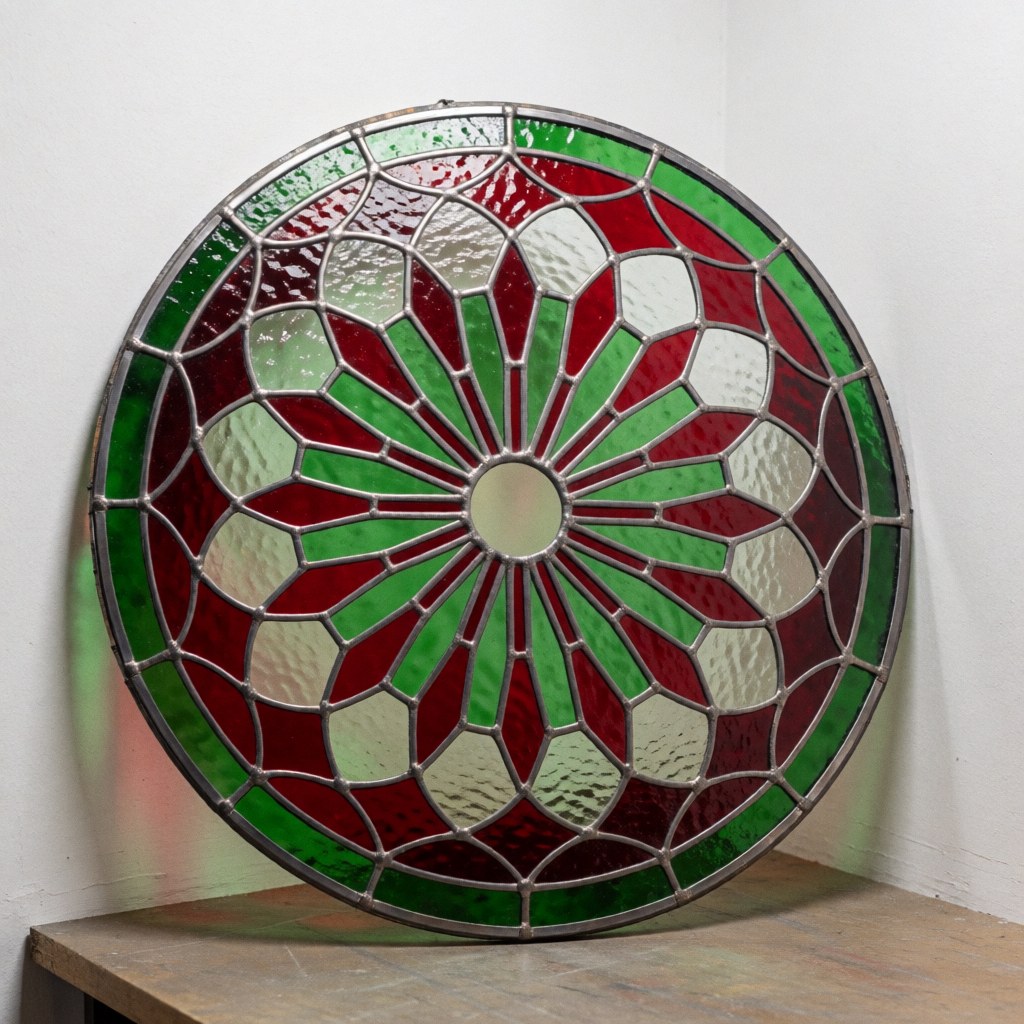 Gros plan sur un oculus en verre coloré avec des motifs radiaux rouges et verts, monté dans un cadre métallique sombre, posé sur une surface blanche.