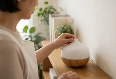 Une thérapeute ajuste un diffuseur sur une étagère en bois, entourée de plantes et baignée de lumière naturelle.