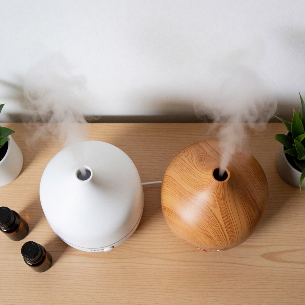 Deux diffuseurs d'aromathérapie, un blanc et l'autre en bois, émettant une vapeur douce sur une table claire, avec des bouteilles d'huile essentielle et des plantes vertes.