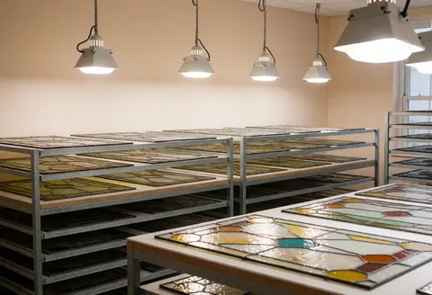 Panneaux de verre décoratif patiné disposés sur des étagères métalliques sous un éclairage industriel dans un atelier propre et organisé.