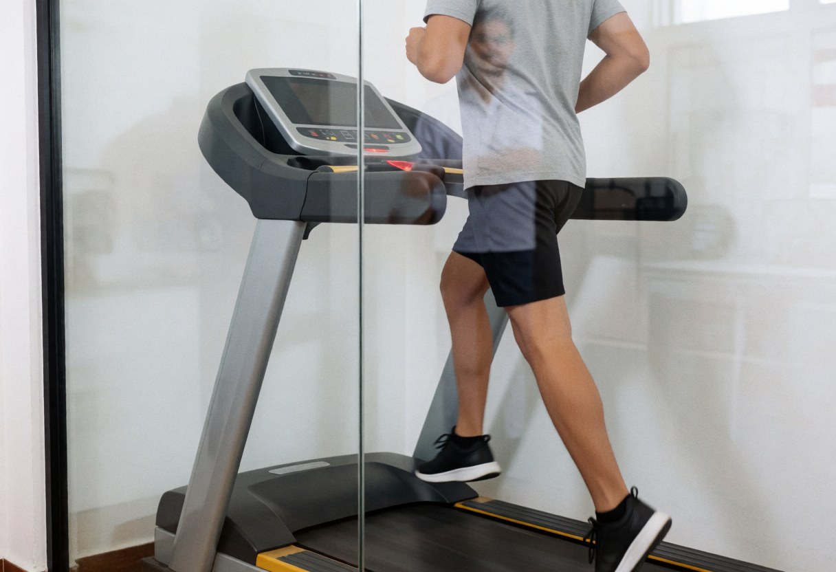 Un athlète effectue un exercice d'entraînement sur un tapis roulant en salle de kinésithérapie, tandis que le reflet d'un thérapeute est visible à travers la vitre transparente.